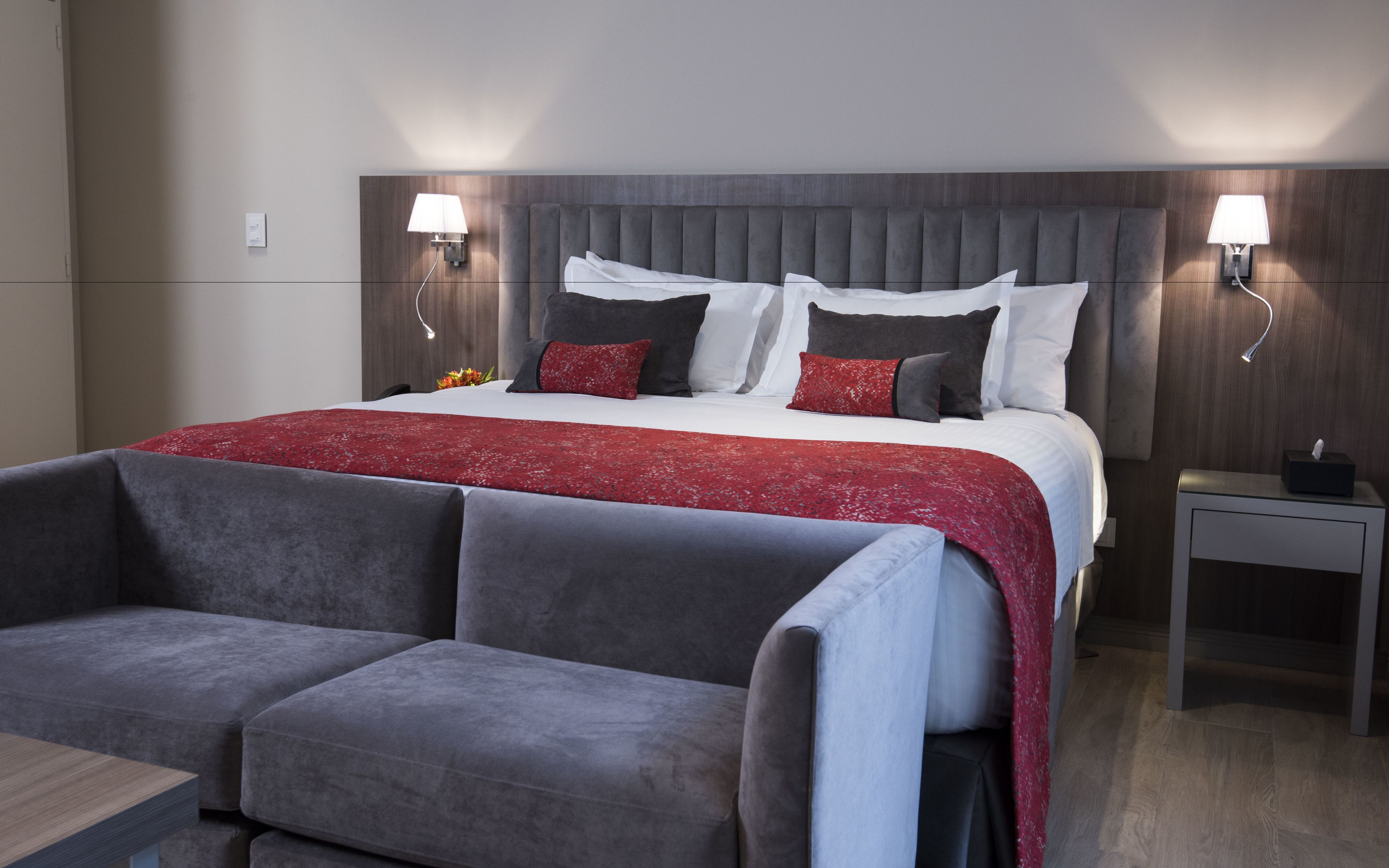 A modern hotel room with a large bed, grey upholstered chairs, a bedside table with a lamp, and a small table with a tissue box.