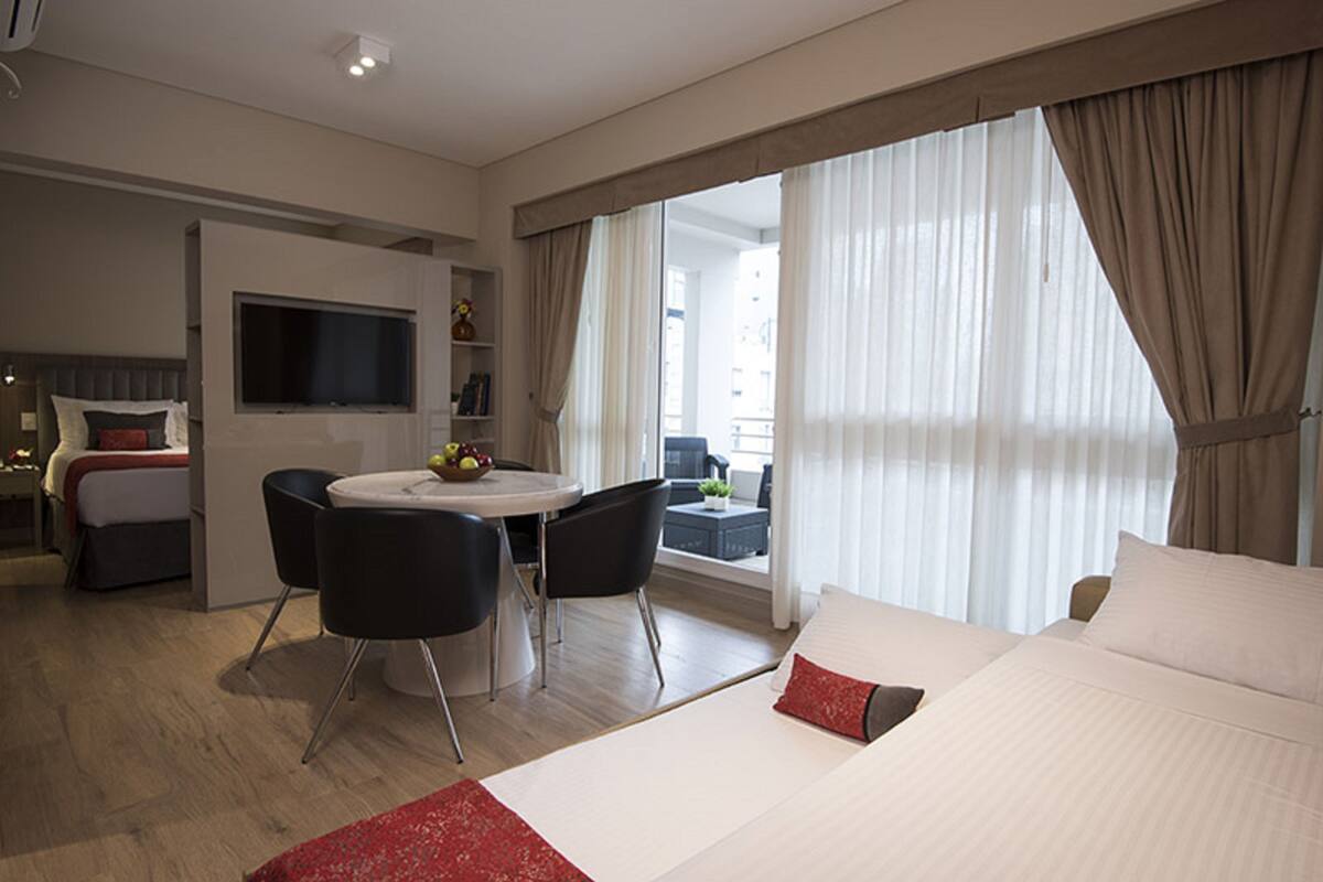 Habitación de hotel con cama, televisor, mesa de comedor con sillas y balcón con cortinas.