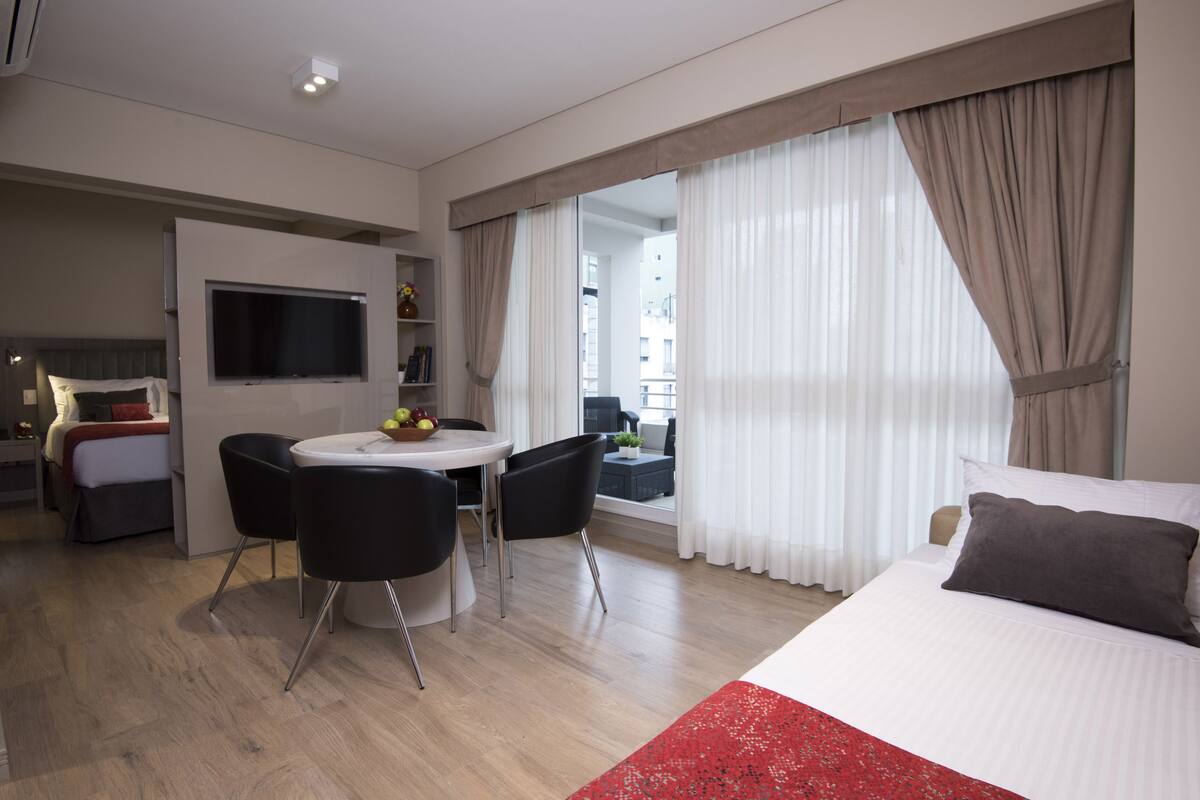 Una habitación de hotel moderna con una cama, un televisor montado en la pared, una zona de comedor con mesa y sillas, y ventanas amplias con cortinas.