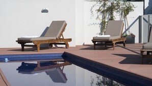 Dos sillones de madera con cojines junto a una piscina, una planta en maceta y un cielo azul despejado.
