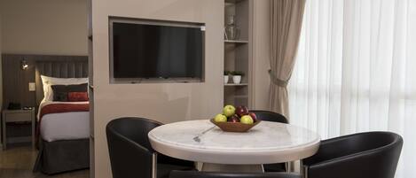 Une chambre d’hôtel moderne équipée d’un lit, d’une télévision, d’une table à manger avec des chaises et d’un panier de fruits.