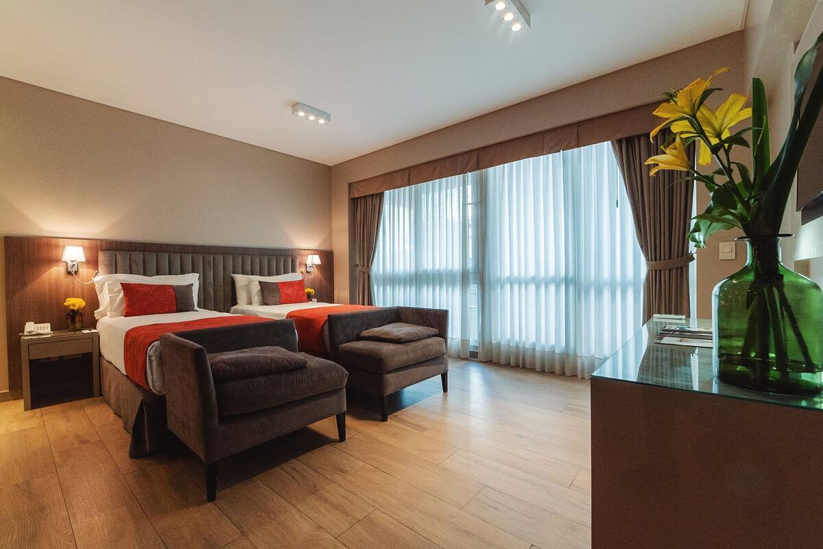 Habitación de hotel con una cama, dos sillones, una mesita de noche, un escritorio y un ventanal con cortinas.