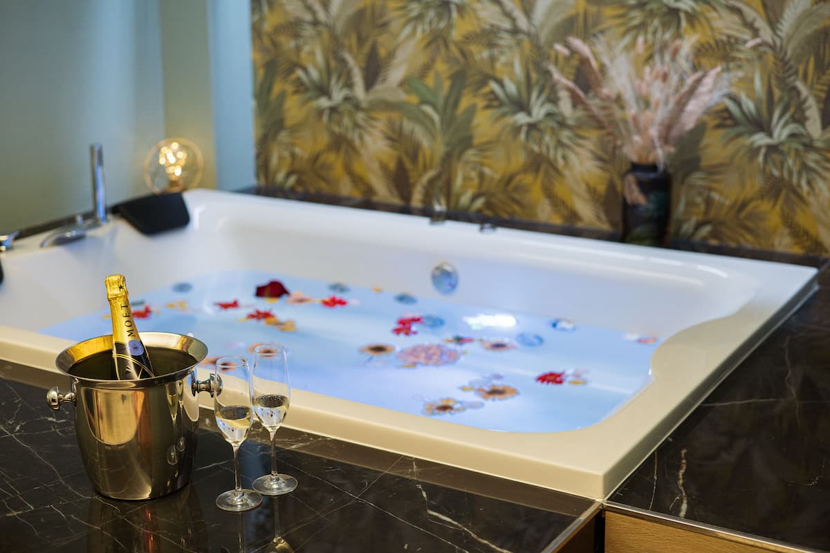 A bathtub filled with water and floral decorations, a champagne bottle, and two glasses on a marble countertop.
