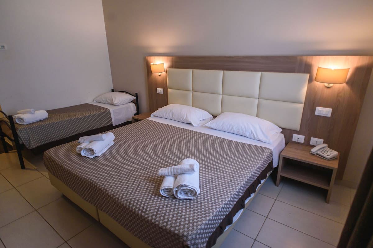 A hotel room with two single beds and a larger bed. Each bed has a pillow and a rolled towel. There is a nightstand with a phone on it.