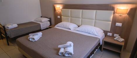 Habitación de hotel con dos camas individuales y una cama más grande. Cada cama tiene un almohadón y una toalla enrollada. Hay una mesita de noche con un teléfono encima.