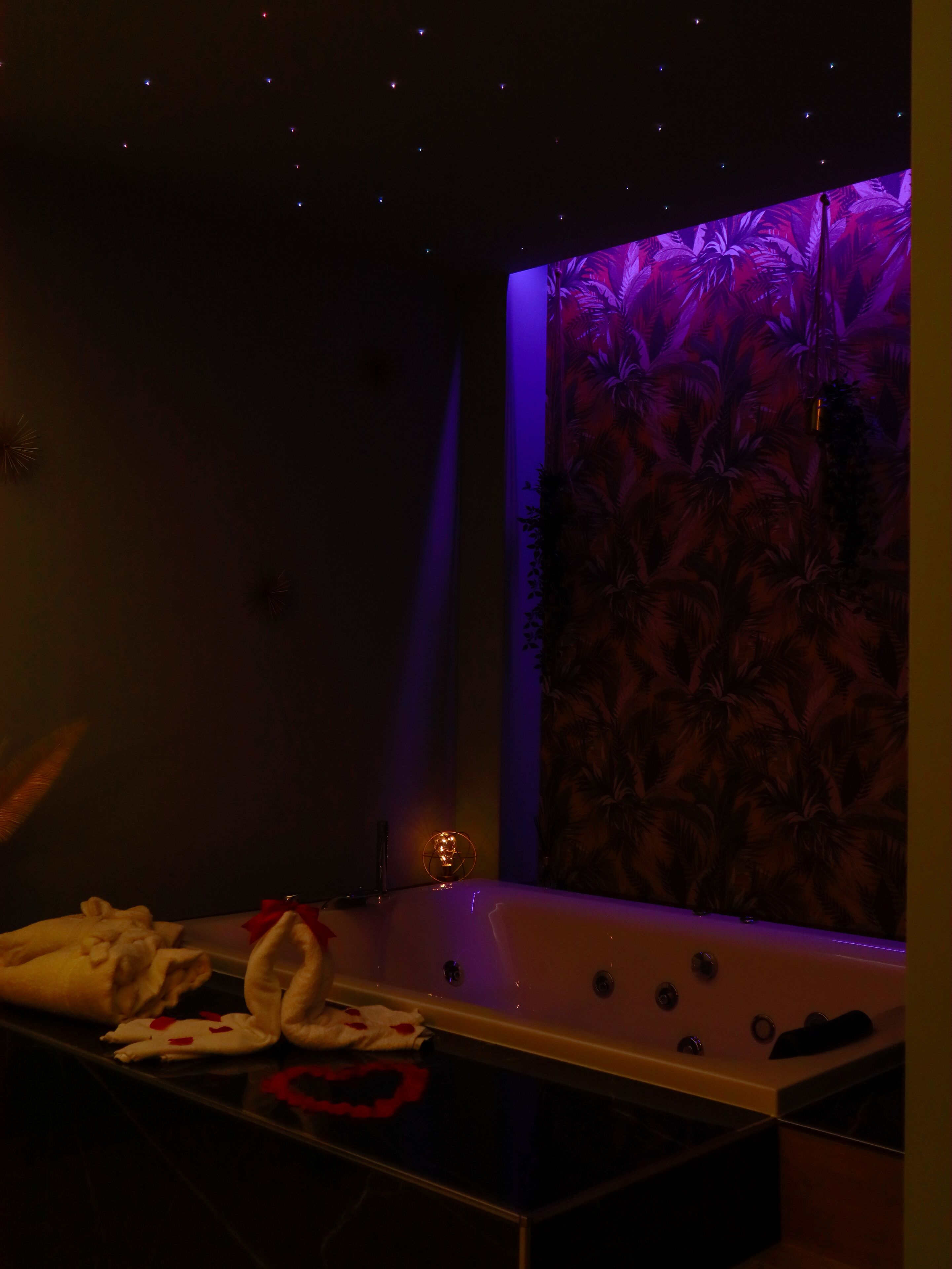 A dimly lit room with a large bathtub, a wall with a tropical leaf pattern, and a small table with towels and a decorative item.