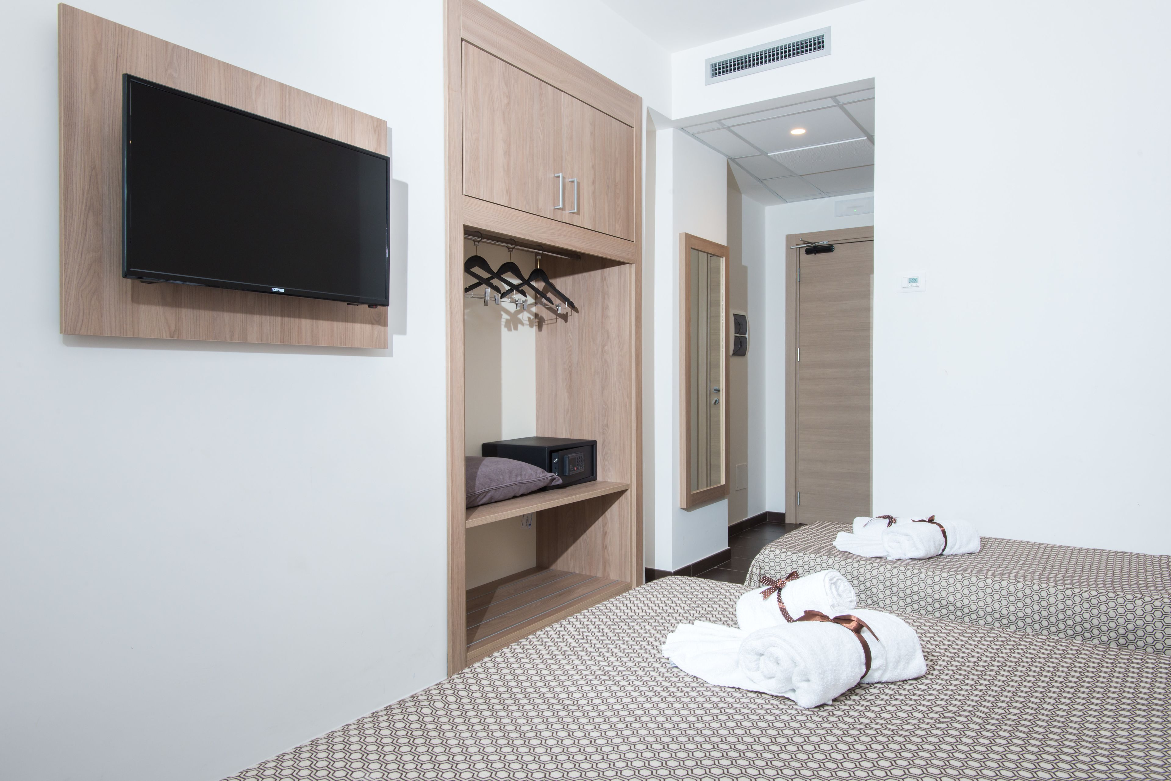 A hotel room with a flat-screen TV mounted on the wall, a wooden wardrobe, a bed with patterned bedding, and two rolled towels on the floor.