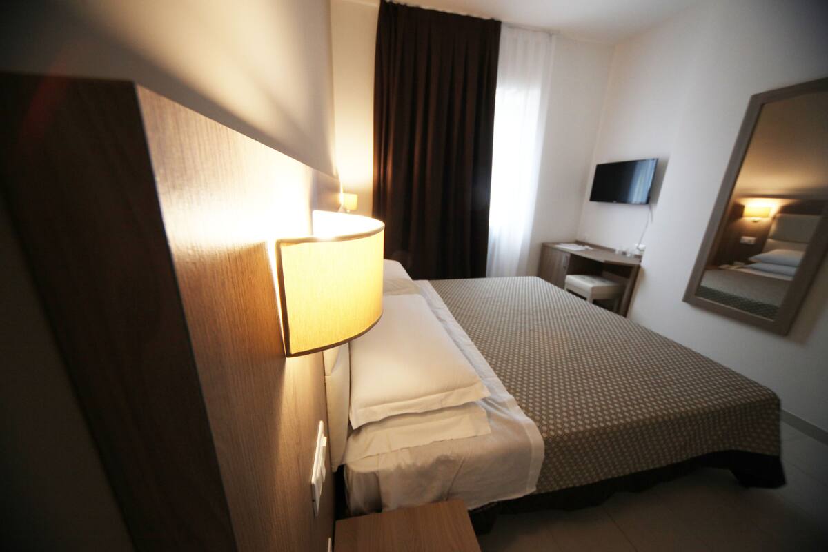 A hotel room with a bed, a bedside table, a lamp, a TV, and a mirror on the wall.