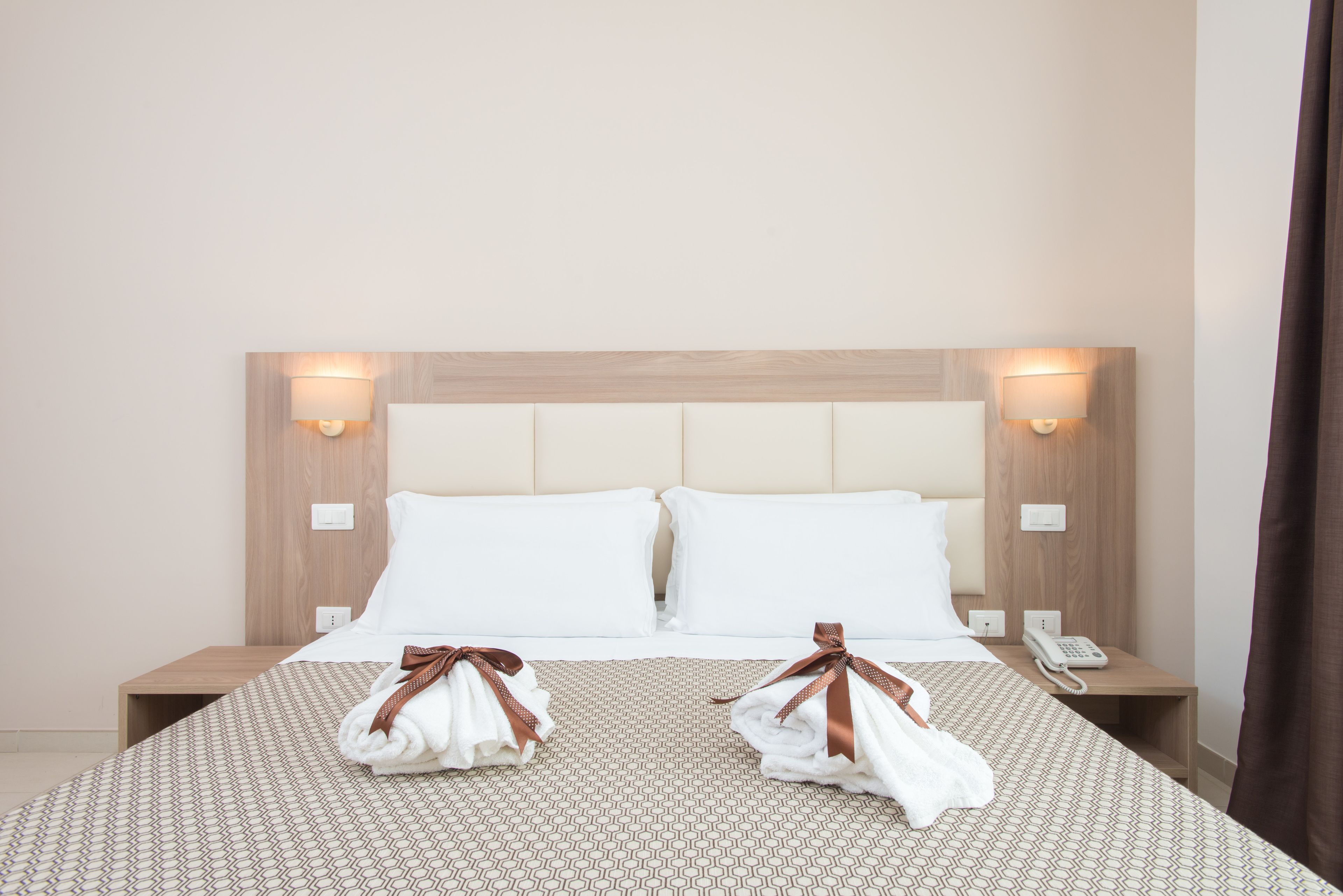 A hotel room with a bed, two pillows, a bedside table with a phone, and a wall-mounted lamp.