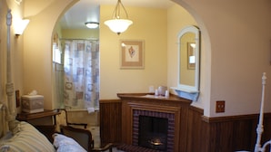 A bedroom with a fireplace, a bed, a chair, and a bathroom visible through an archway.