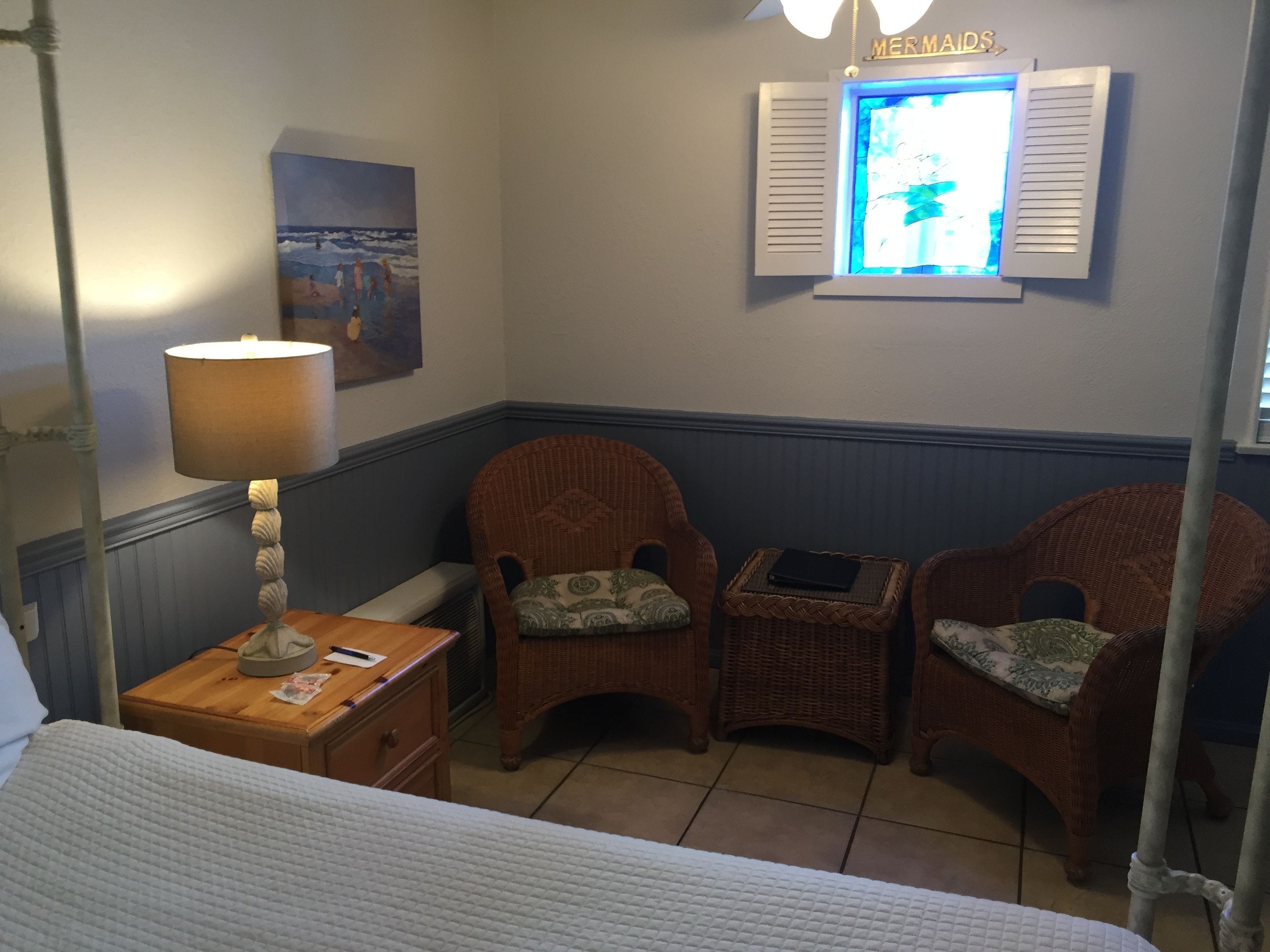 A room with a bed, a nightstand, two wicker chairs, a small table with a lamp, a painting on the wall, and a window with shutters.