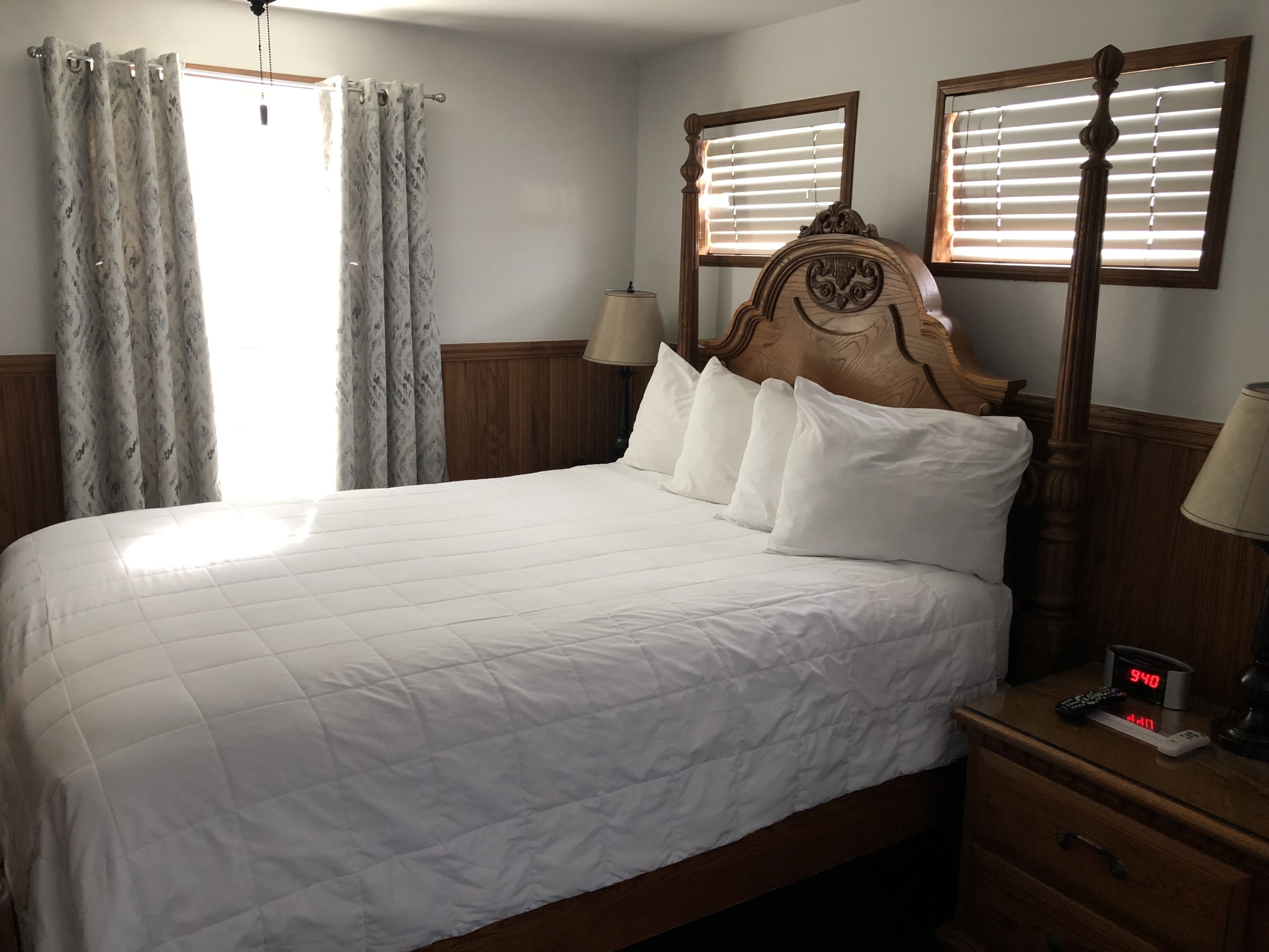 Una cama bien hecha con tablero de madera, dos lámparas de noche, una mesita con un reloj digital y una ventana con persianas.