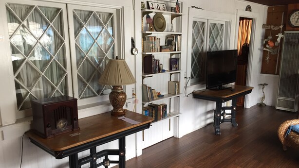 A room with wooden flooring, a vintage radio, a lamp, a television on a wooden stand, and a bookshelf filled with books and decorative items.
