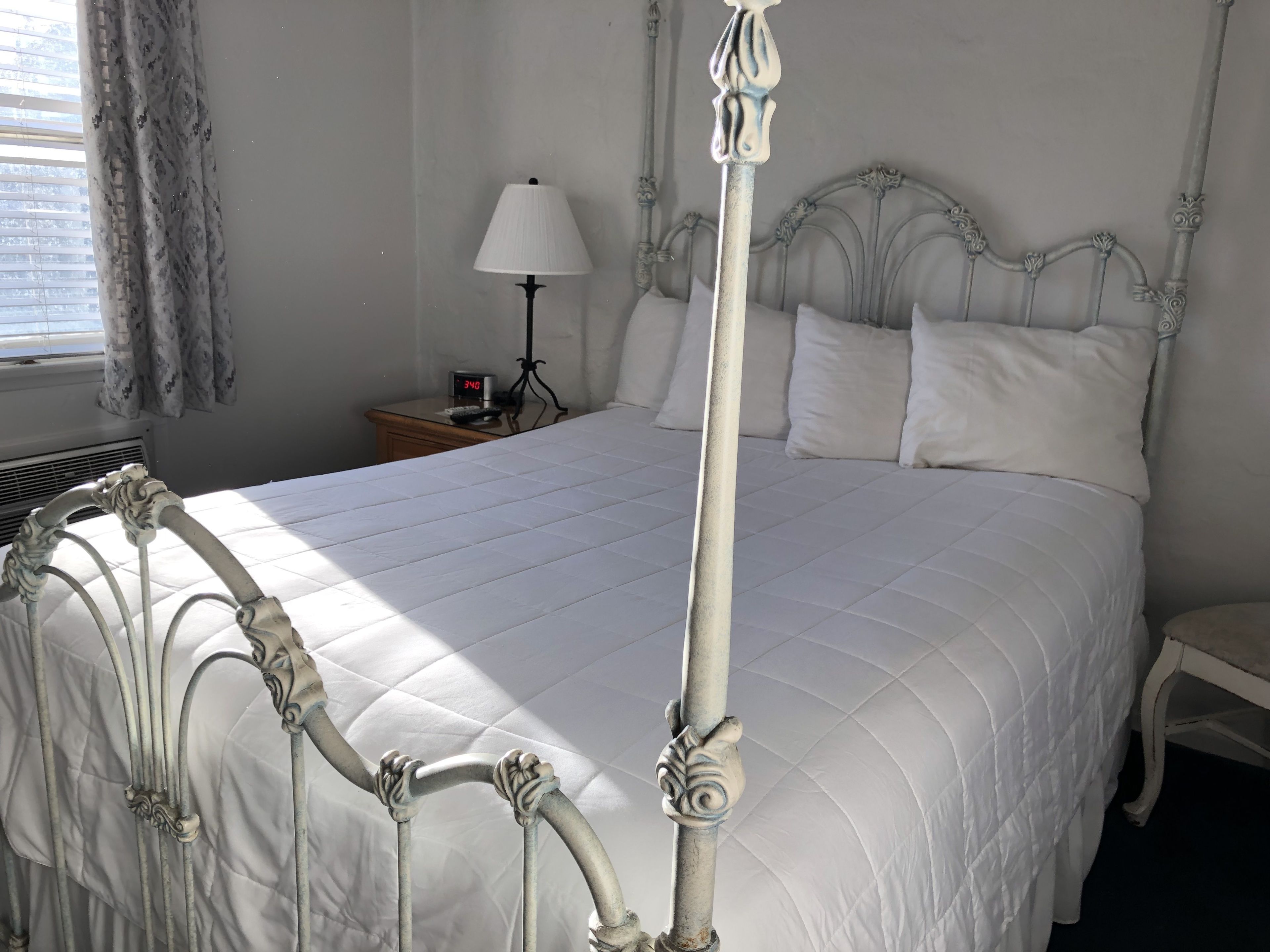 A white four-poster bed with a quilted mattress, a bedside table with a lamp, and a clock displaying 3:40.