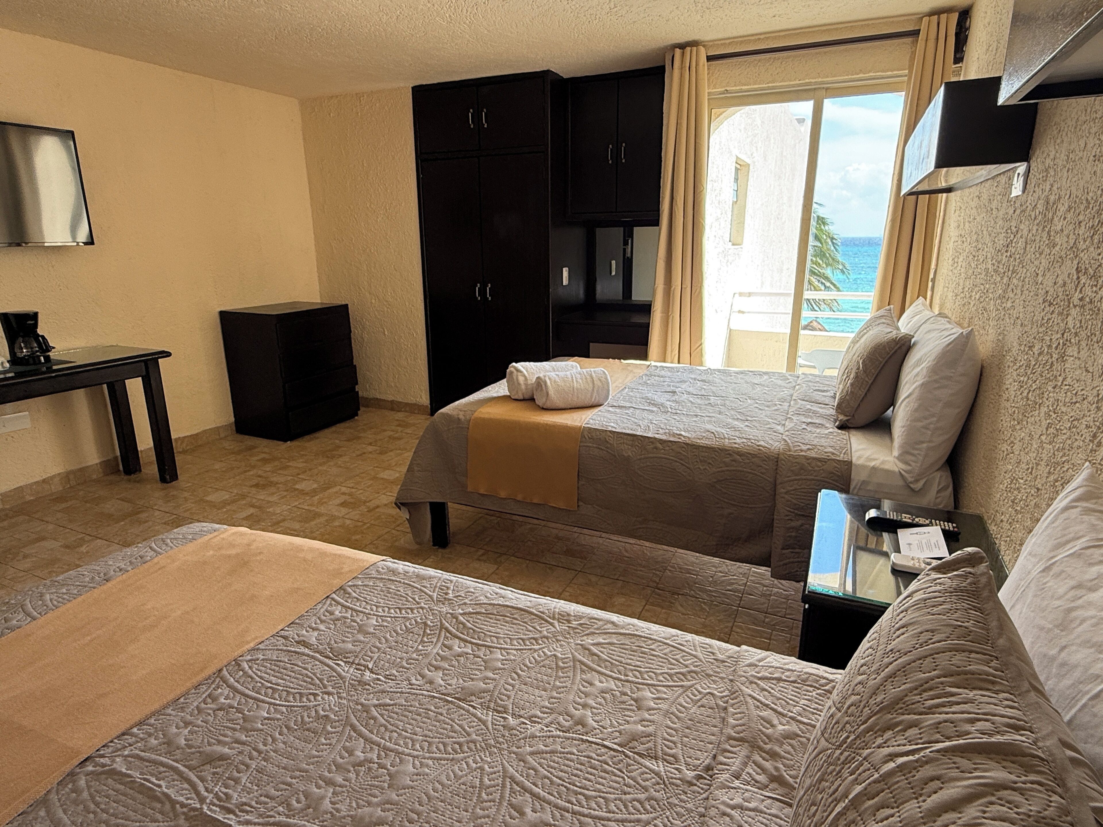 A hotel room with two beds, a desk, a wardrobe, and a view of the sea.