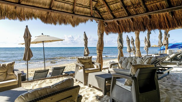 Un área de descanso en la playa con mobiliario de mimbre, sombrillas y techo de paja.