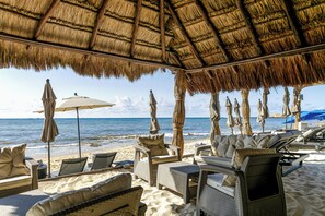 A beachside lounge with wicker furniture, umbrellas, and a thatched roof.