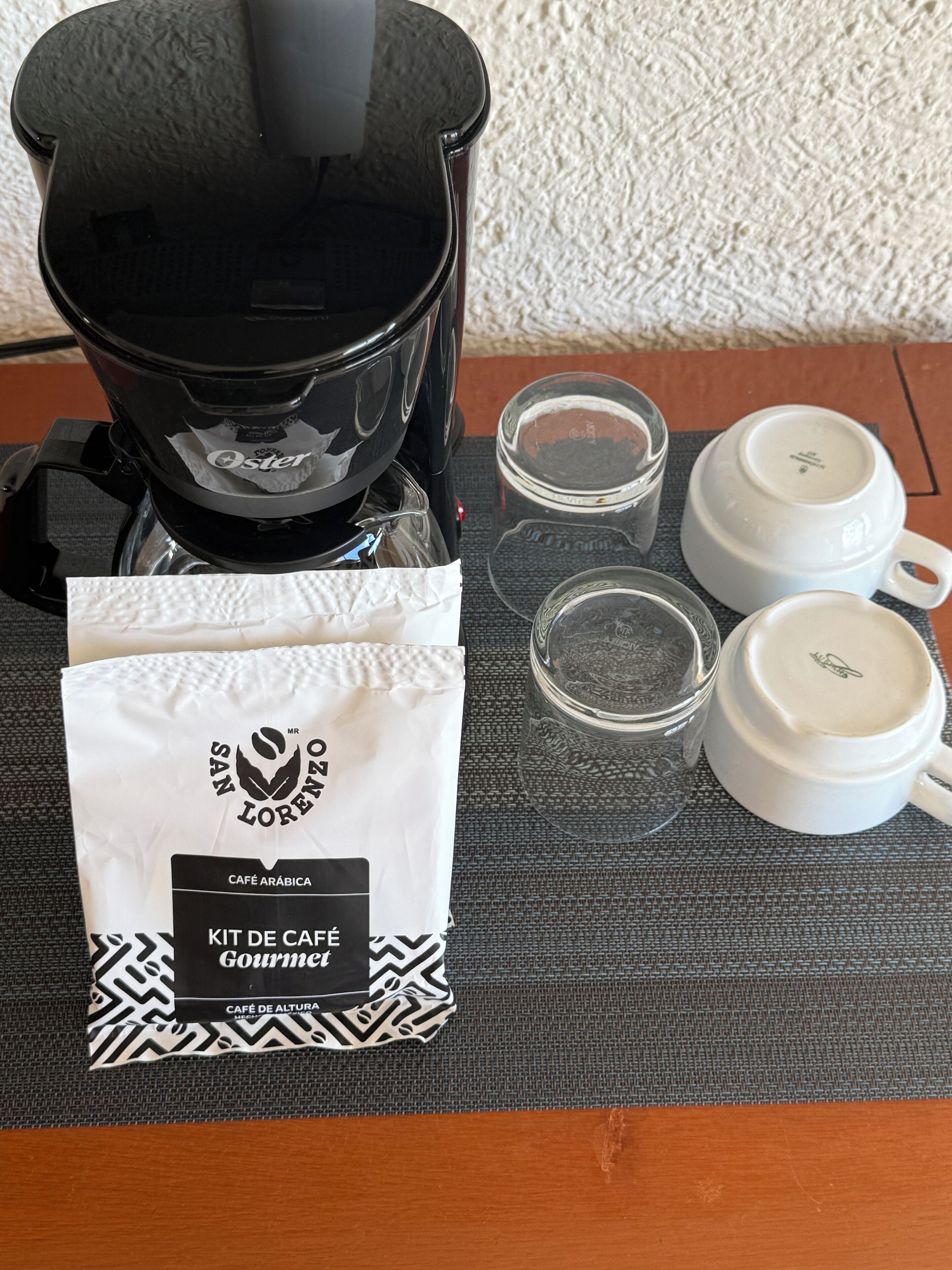 A coffee maker, a white bag with a logo, two white cups, and a glass on a table.