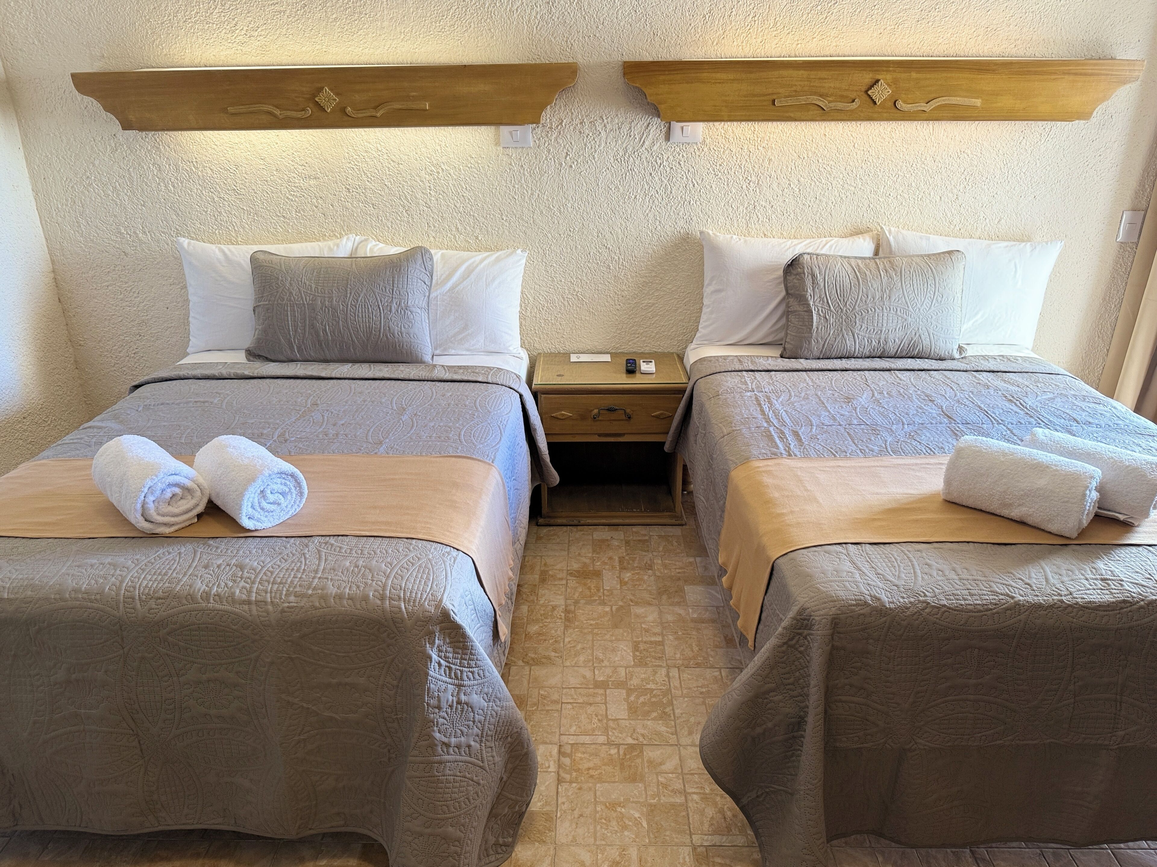 A hotel room with two beds, each with a wooden headboard, a nightstand, and a small drawer.