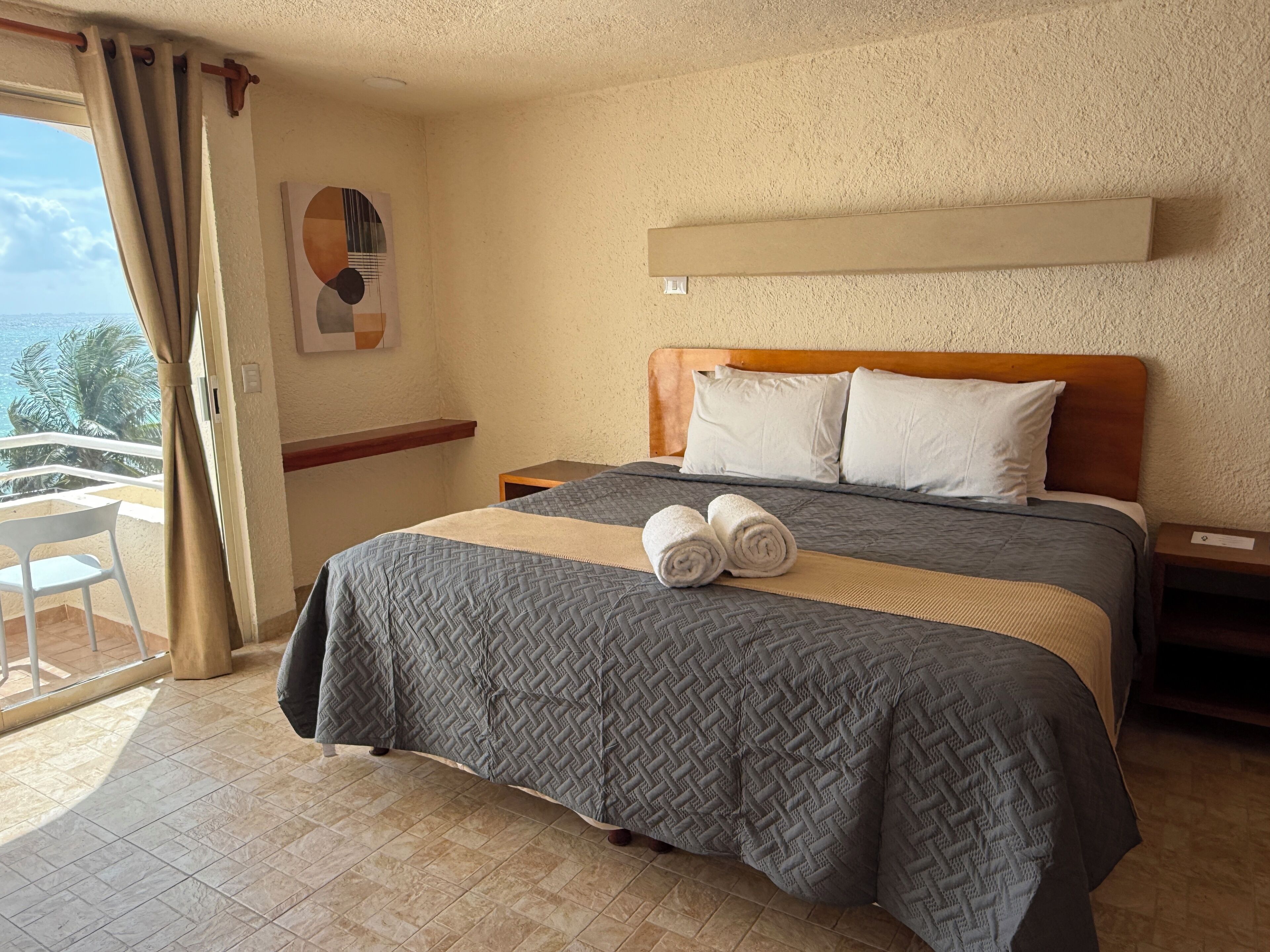 Ein Hotelzimmer mit Bett, Holz-Kopfteil, zwei weiße Kissen, einem beigen Nachttisch, einem kleinen Tisch mit Stuhl und Meerblick durch ein mit Vorhängen versehenes Fenster.