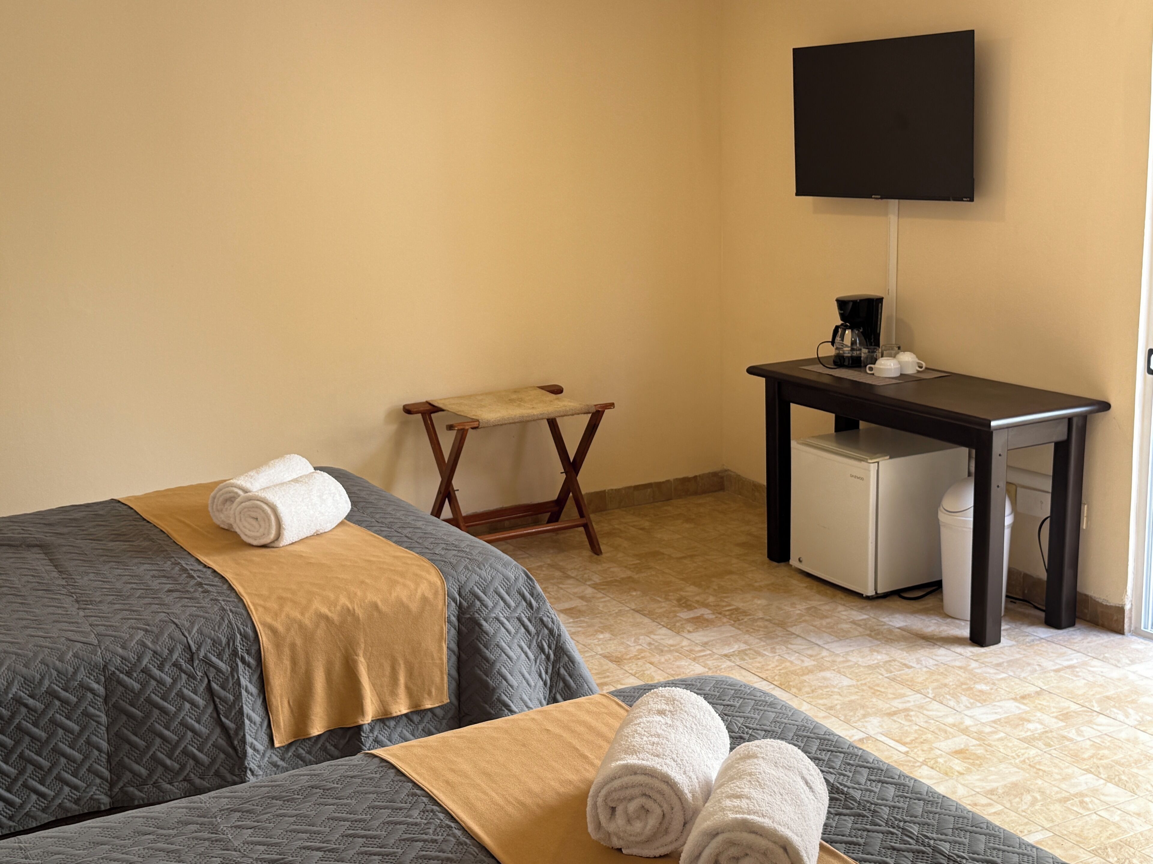 A hotel room with two beds, a small table with a coffee maker, a TV on the wall, and a compact refrigerator.