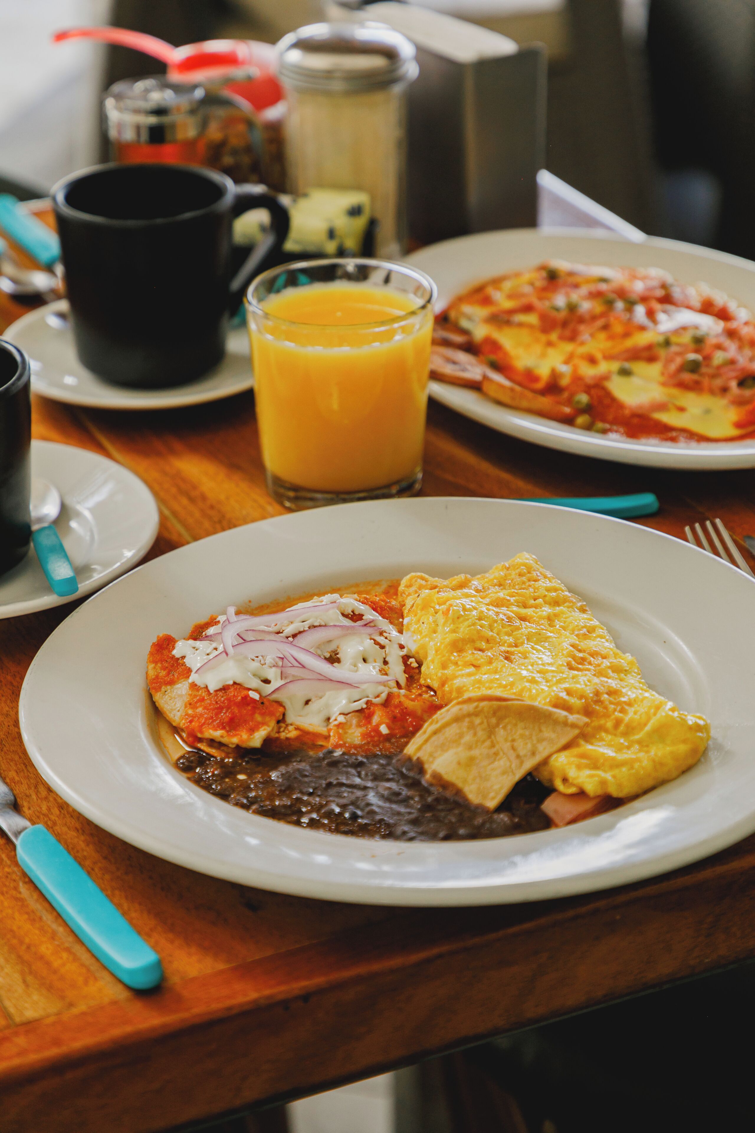 Prato de café da manhã com ovos, queijo e molho de tomate, um copo de suco de laranja e uma xícara de café sobre uma mesa de madeira.