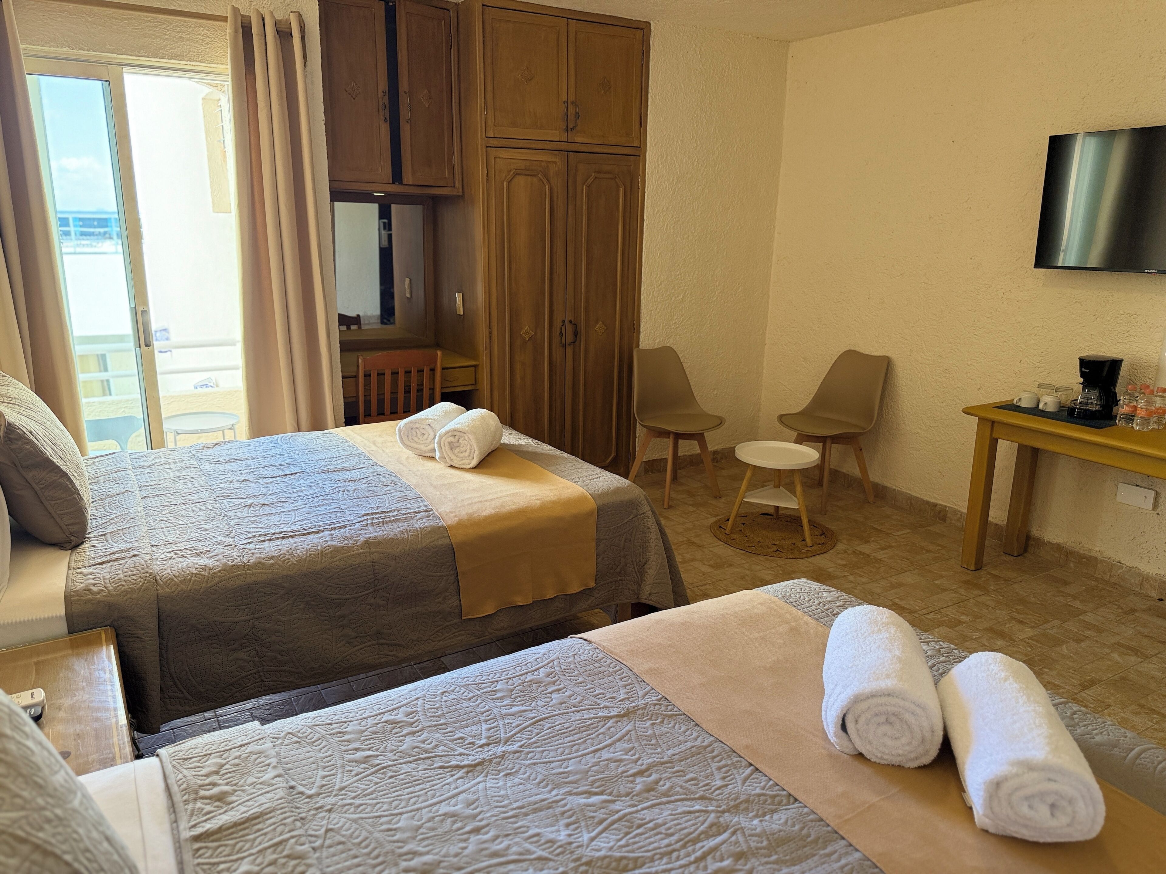 A hotel room with two beds, a desk, chairs, a TV, and a balcony with curtains.