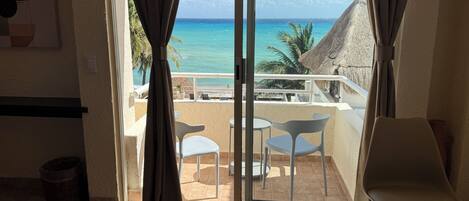 A room with a view of the ocean, a balcony with chairs, and a thatched roof structure.