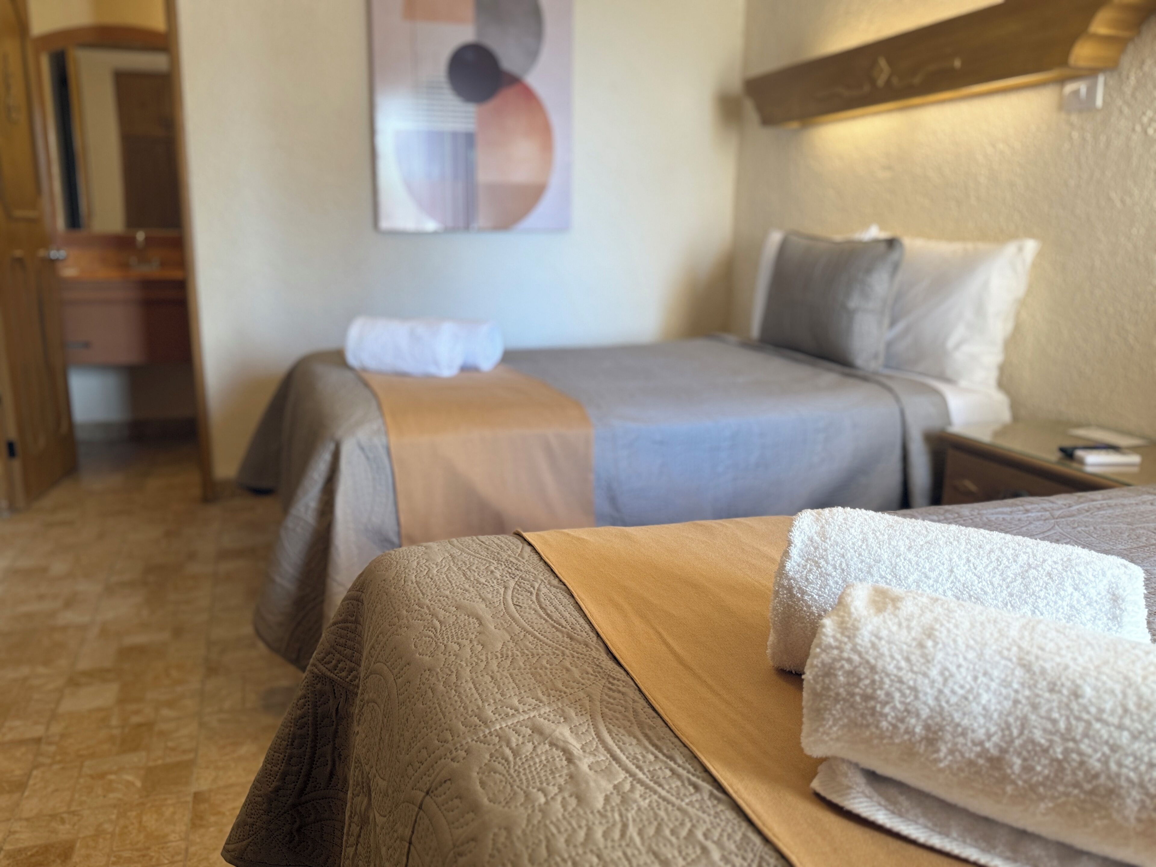 A hotel room with two beds, a wooden headboard, a painting on the wall, and a nightstand with a lamp.