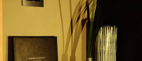 A room with a wall-mounted clock, a framed certificate, a vase with green plants, and a book on a table.