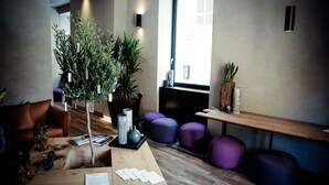 Et moderne loungeområde med et træbord, en potteplante, lilla siddepladser og et vindue med naturligt lys.