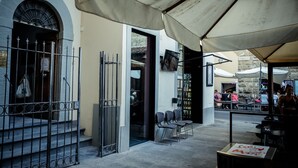 En udendørs café med borde og stole, et overdækket siddeområde og en bygningseingang med en metalport.