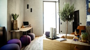 Et moderne interiør med et træskrivebord, lilla siddepladser, en stor plante og udsigt gennem en glasdør.