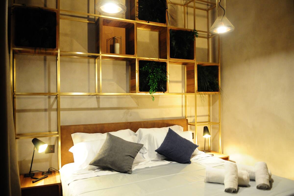 A neatly made bed with pillows, a bedside lamp, and a wall-mounted shelf with a plant.
