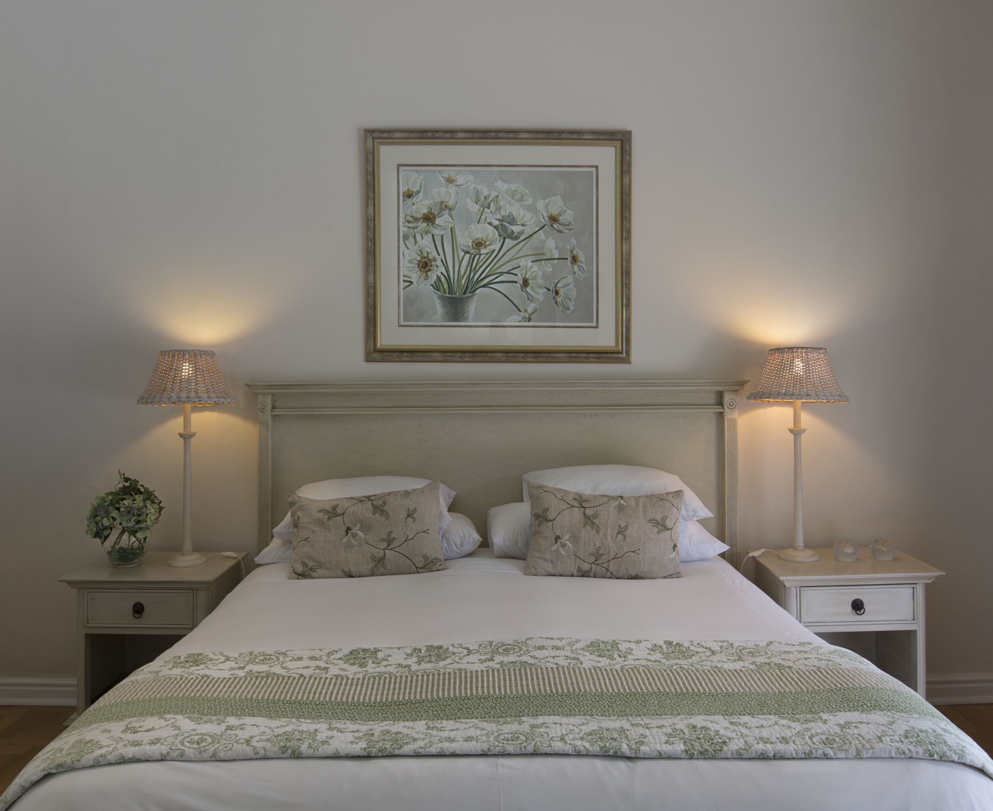 Cama bem arrumada com roupa de cama floral, duas luminárias de cabeceira e uma obra de arte floral emoldurada acima da cama.