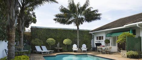 Una piscina con sillones de descanso y una mesa, rodeada de vegetación y una casa con tejado de baldosas.