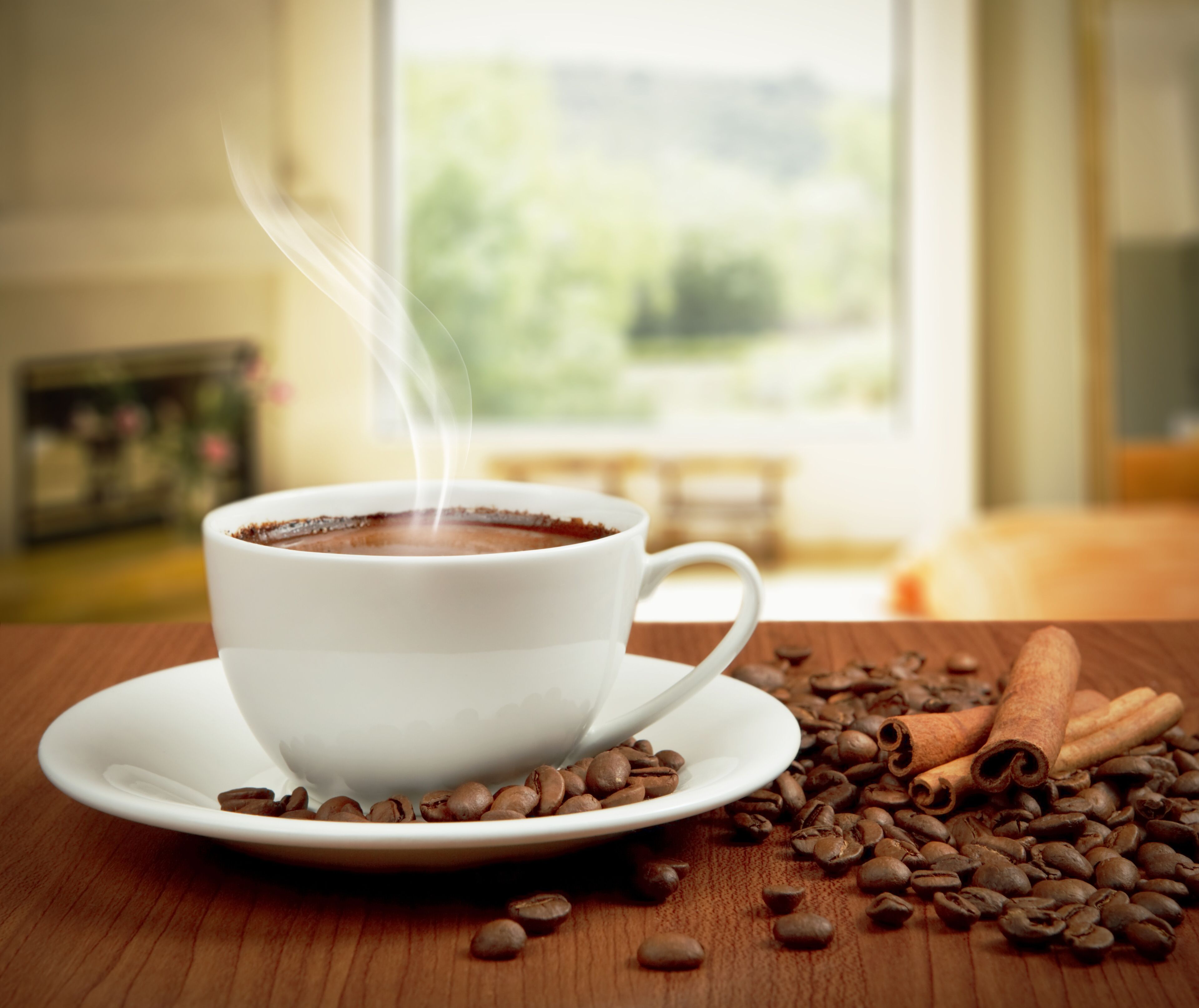 A white cup of coffee on a saucer with steam rising, surrounded by coffee beans and cinnamon sticks on a wooden table.