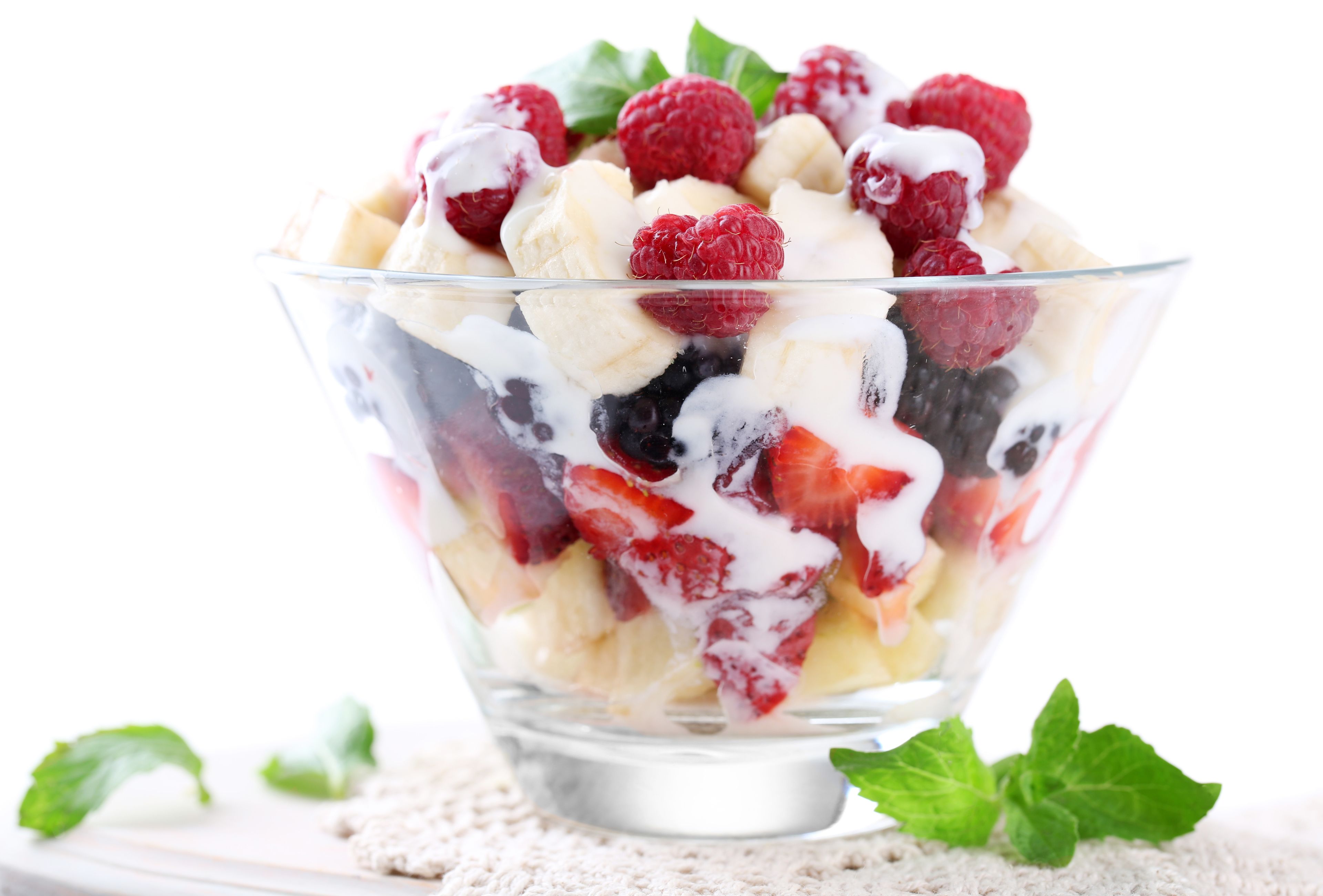 Un saladier en verre transparent rempli d’une salade de fruits, comprenant des fraises, des framboises et des bananes, le tout surmonté d’une cuillère de crème.