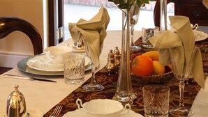 Mesa de jantar posta com pratos brancos, talheres e copos, com um vaso de rosas vermelhas e uma tigela de frutas.