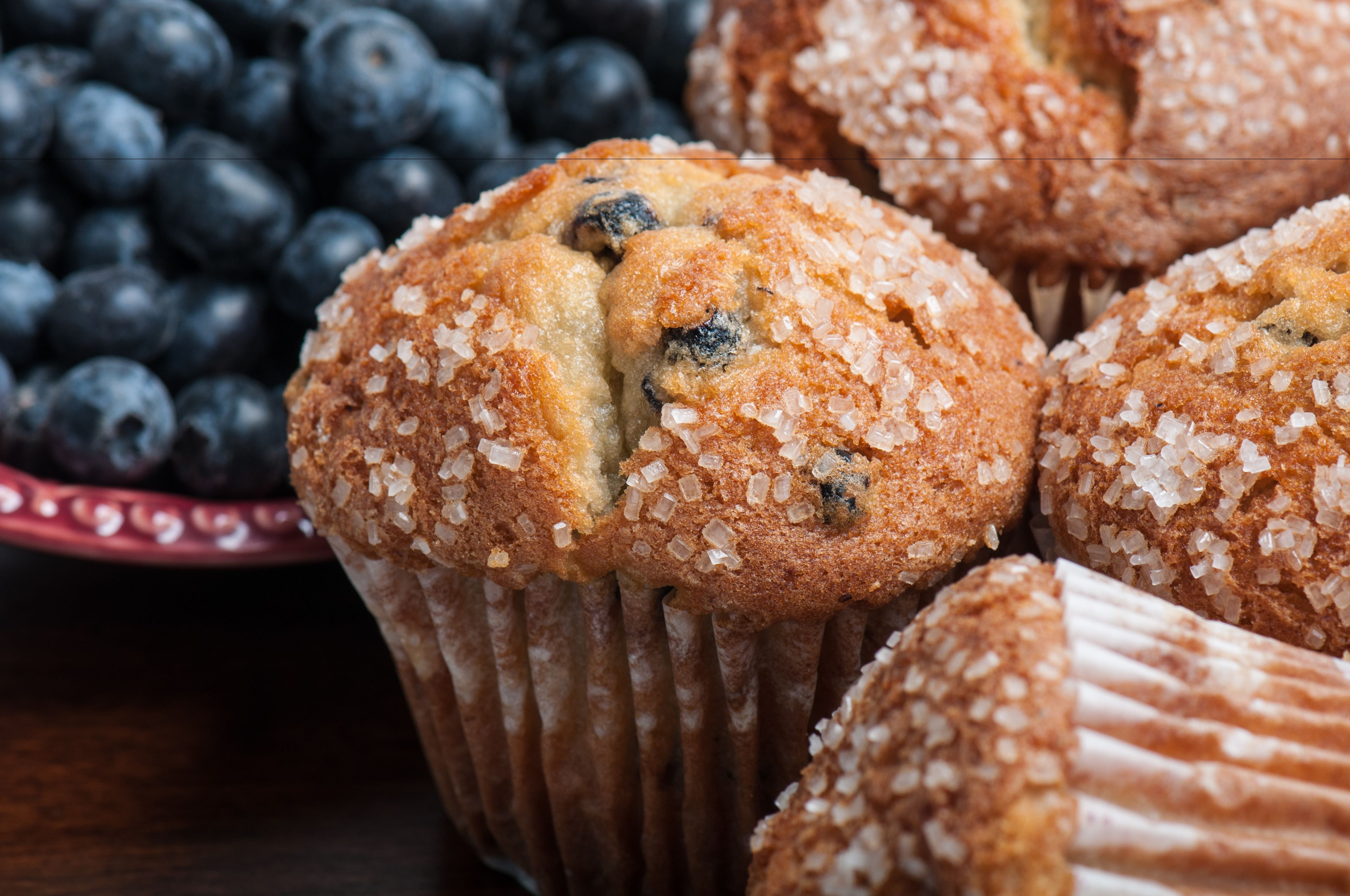 Un muffin con mirtilli e zucchero sopra, posizionato su un piatto con una ciotola di mirtilli sullo sfondo.