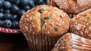 Un muffin con mirtilli e zucchero sopra, posizionato su un piatto con una ciotola di mirtilli sullo sfondo.