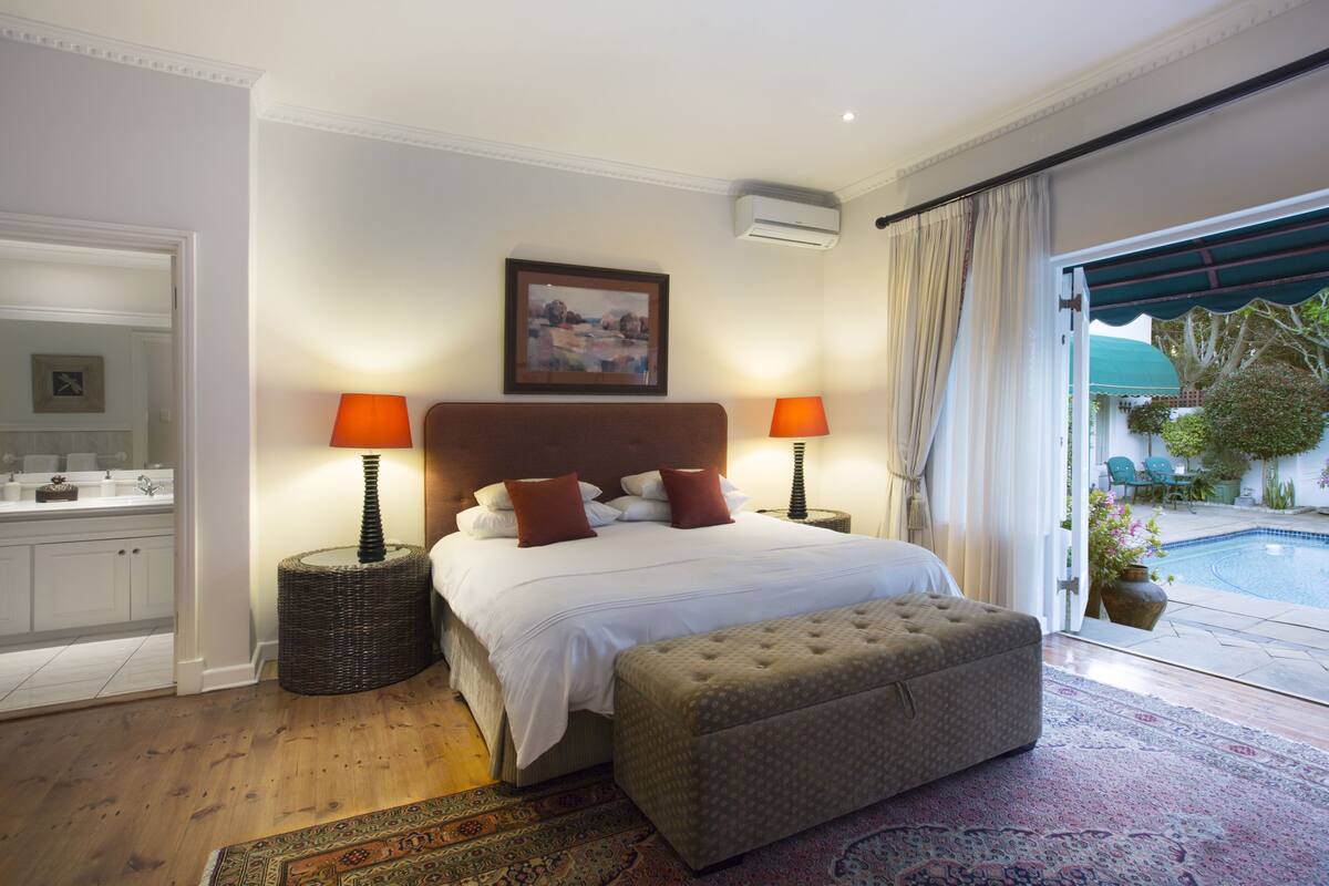A bedroom with a large bed, two bedside lamps, a wicker side table, a bench, a painting on the wall, and a view of a swimming pool through a glass door.