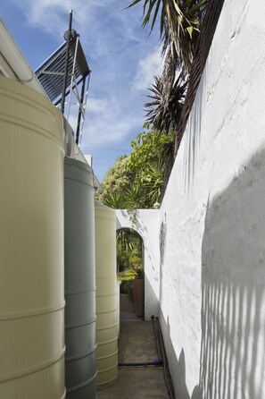 A narrow alleyway with water tanks on one side and a white wall on the other, leading to a covered area with plants. - Carslogie House (Gqeberha (Port Elizabeth))