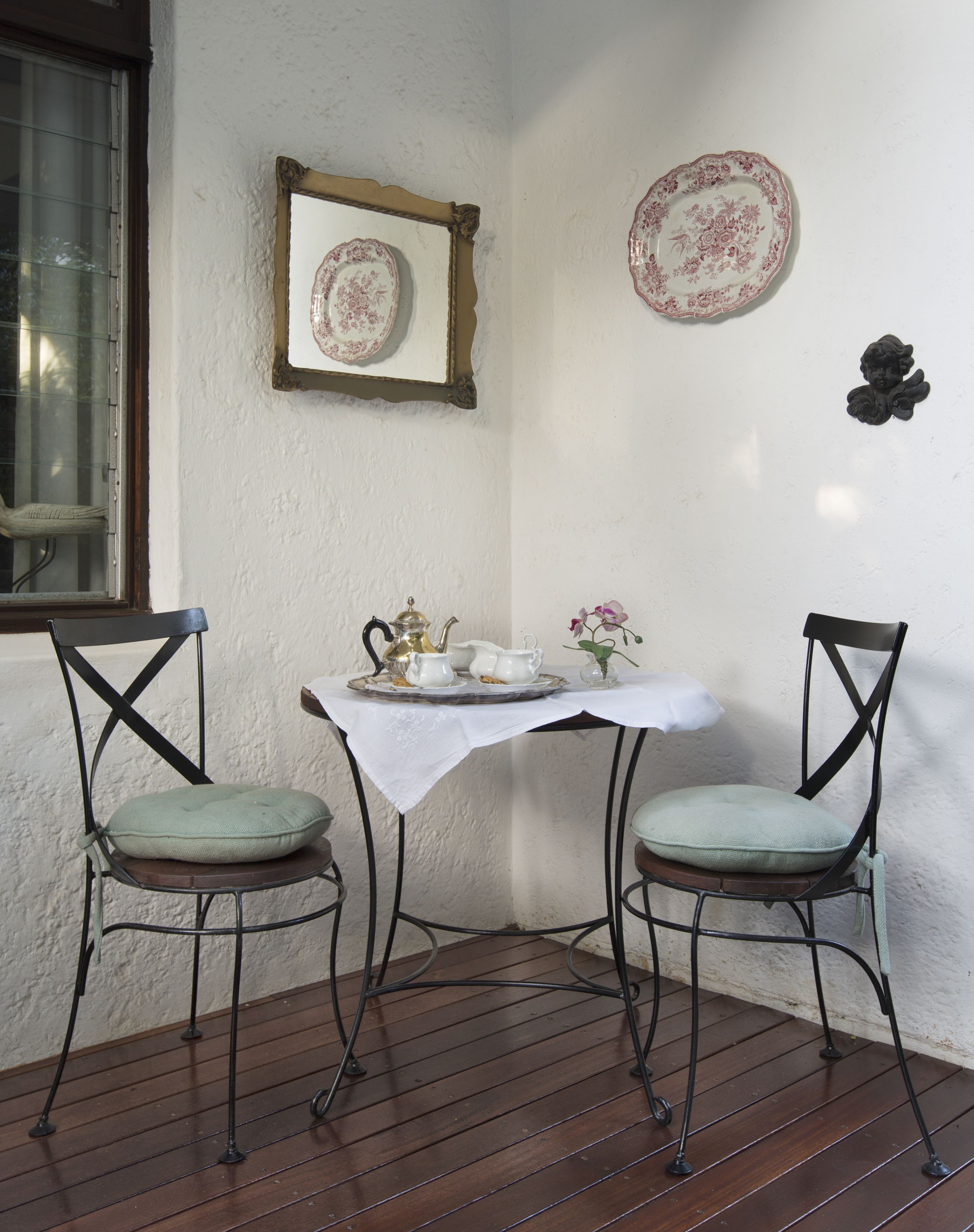A small, enclosed patio area with a table set for tea, two chairs, a decorative mirror, and wall-mounted plates.