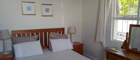A bedroom with a wooden headboard, a bed with white and beige bedding, two bedside tables with lamps, a dresser, and a window with curtains.