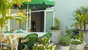 Een terras buiten met een tafel en stoelen, omringd door potplanten en een groene parasol.