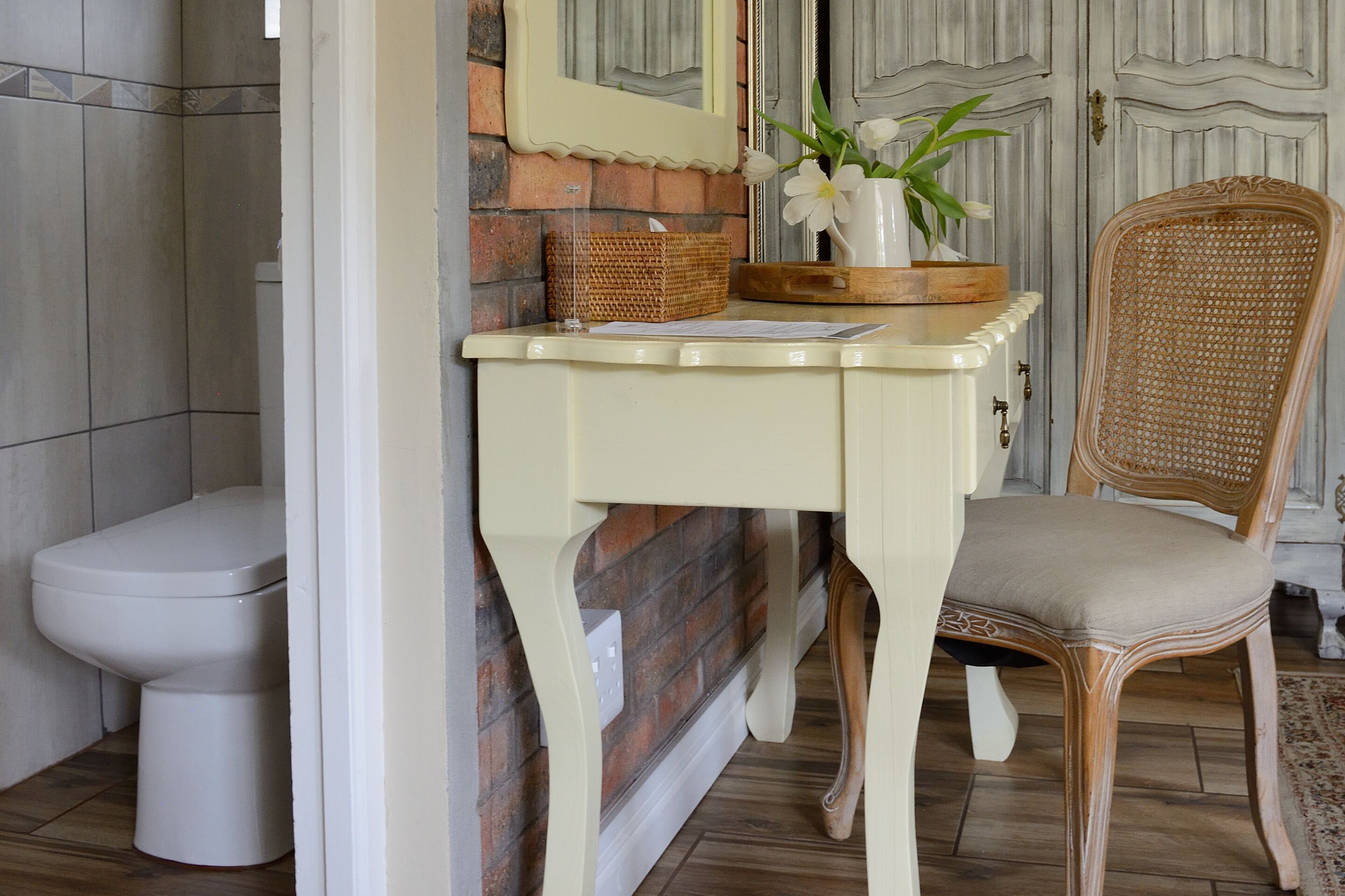 A bathroom with a toilet, a wicker chair, a small table with a vase of flowers, and a mirror on the wall.