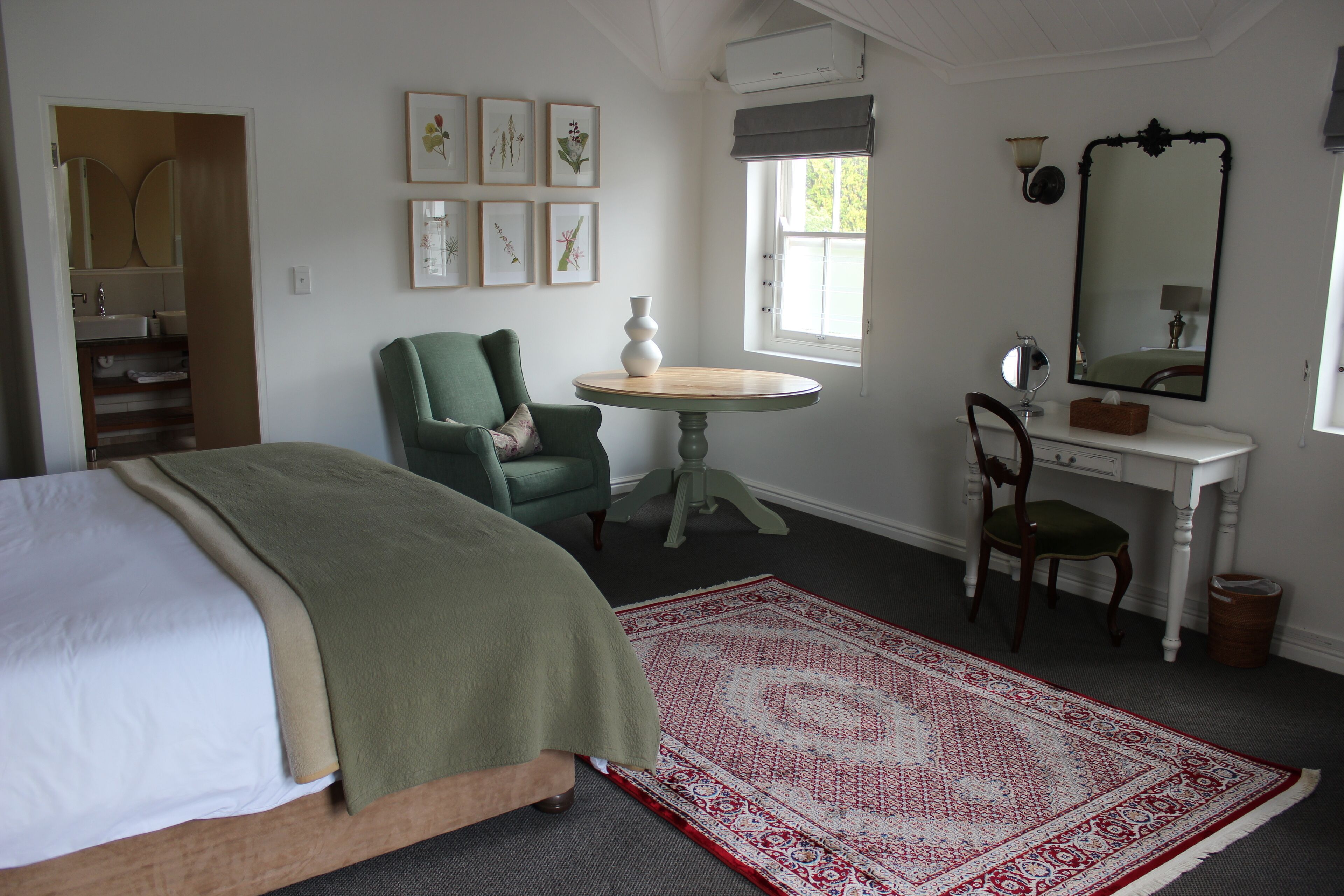 A bedroom with a bed, a green armchair, a desk with a chair, a round table, and a large mirror.
