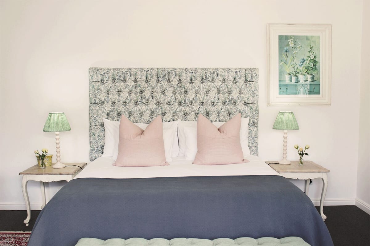 A bedroom with a patterned headboard, two bedside tables with lamps, and a framed floral painting on the wall.