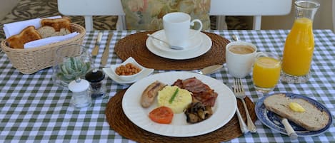 Una mesa de desayuno con mantel a cuadros, una cesta de pan, un plato con huevos, tocino, champiñones y un vaso de jugo de naranja.